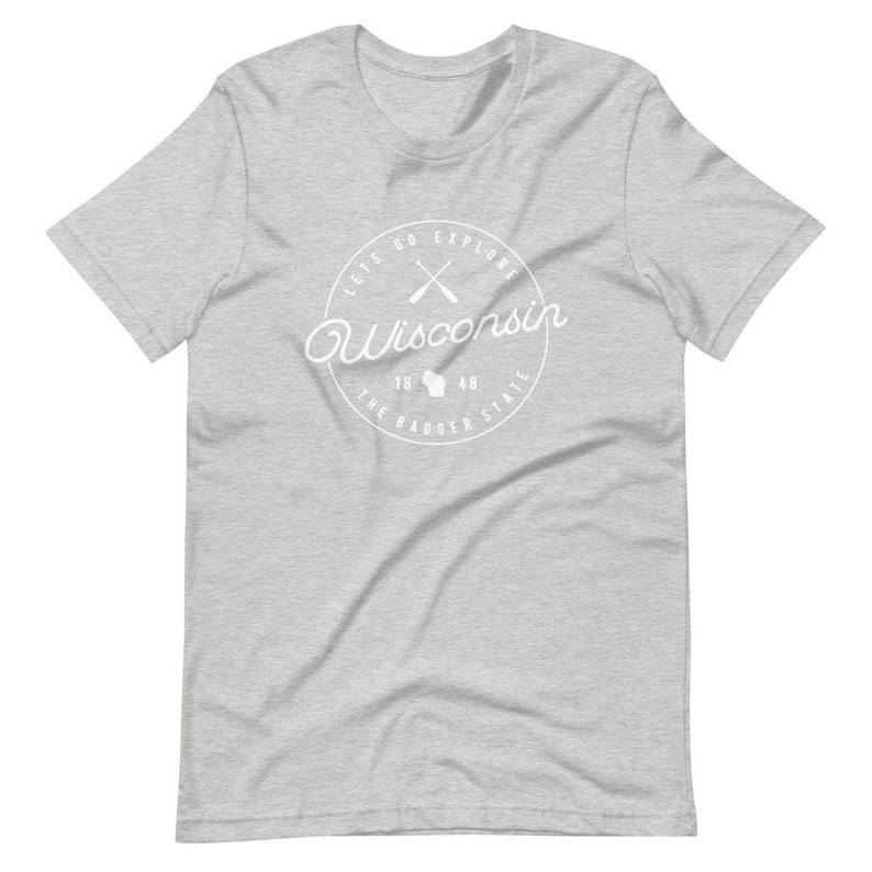 May include: A heather grey t-shirt with a white circle graphic that says "Let's Go Explore Wisconsin" with a white outline of the state of Wisconsin and two crossed paddles inside the circle. The text "The Badger State" is written below the state outline and the years "1848" are written above the state outline.