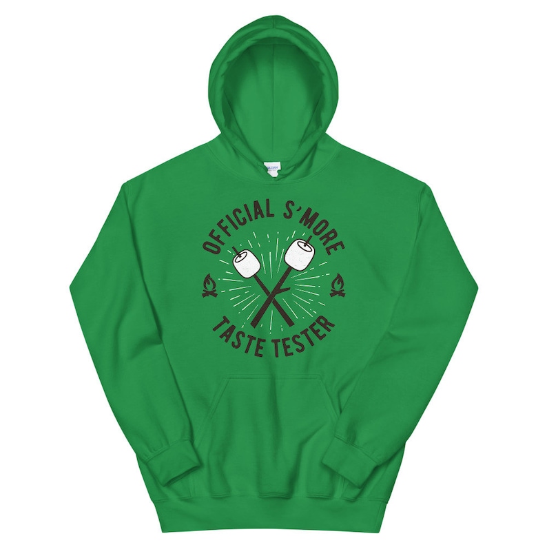 K&ouml;nnte beinhalten: Ein leuchtend gr&uuml;ner Kapuzenpullover mit dem Text "OFFICIAL S'MORE TASTE TESTER" in einem kreisf&ouml;rmigen Design. Das Design umfasst gekreuzte Marshmallow-Sticks und kleine Lagerfeuer-Grafiken. Der Kapuzenpullover hat eine Vordertasche und eine Kapuze mit Kordelzug.