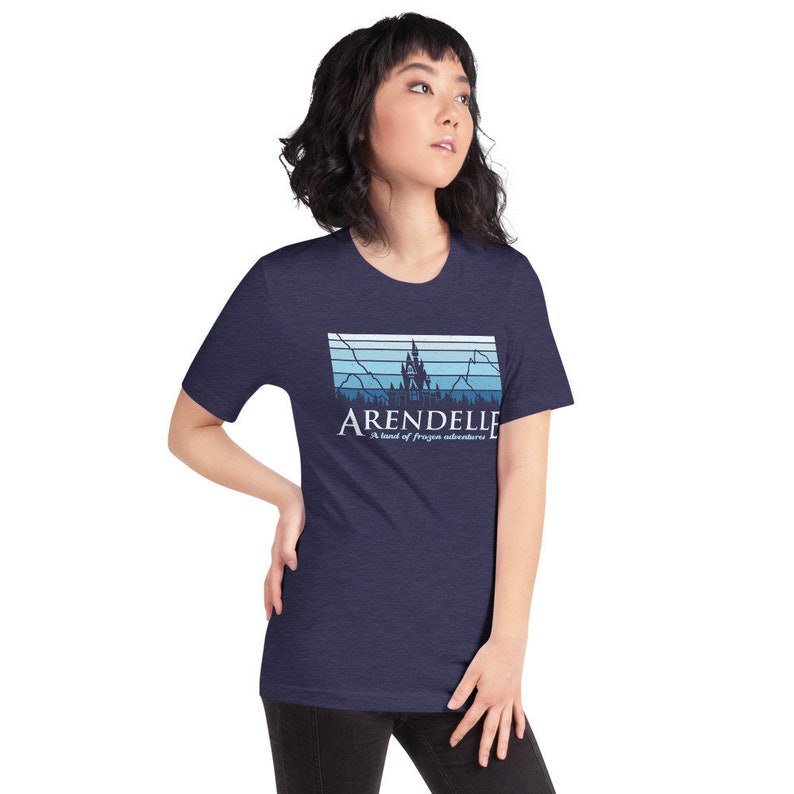 May include: A navy blue t-shirt with a graphic of a castle and mountains in a blue and white color scheme. The text "Arendelle" and "A land of frozen adventures" is printed on the shirt.