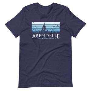 May include: A navy blue t-shirt with a retro-style graphic of a castle with mountains and trees in the background. The text "Arendelle" is printed below the graphic, along with the phrase "A land of frozen adventures".
