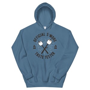 K&ouml;nnte beinhalten: Ein blauer Kapuzenpullover mit dem Text "OFFICIAL S'MORE TASTE TESTER" in einem kreisf&ouml;rmigen Design. Das Design umfasst gekreuzte Marshmallow-St&auml;be und kleine Lagerfeuer-Grafiken. Der Kapuzenpullover hat eine Vordertasche und eine Kapuze mit Kordelzug.