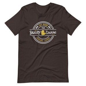 May include: A dark brown t-shirt with a white circle logo that says "The Snuggly Duckling Tap Room Karaoke Lounge" with a yellow rubber duck illustration.