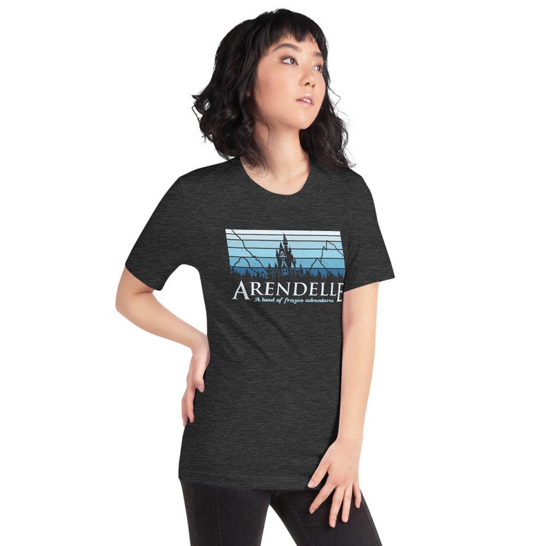 May include: A dark heather grey t-shirt with a retro-style graphic of a castle and mountains. The text "ARENDELLE" is printed in white with the words "A land of frozen adventures" below it.
