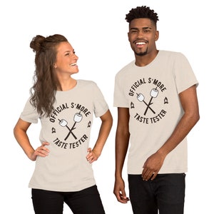 May include: Two people wearing beige t-shirts with a black graphic that says "Official S'more Taste Tester" and a drawing of two marshmallows on sticks crossed over each other.