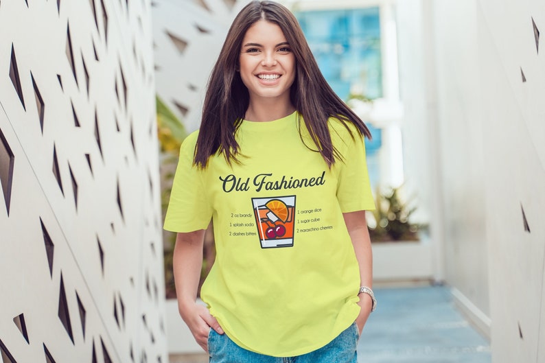 May include: A bright yellow t-shirt with a graphic of an Old Fashioned cocktail recipe. The text on the shirt reads "Old Fashioned" and includes the ingredients: 2 oz bourbon, 1 splash soda, 2 dashes bitters, 1 orange slice, 1 sugar cube, 1 maraschino cherry.