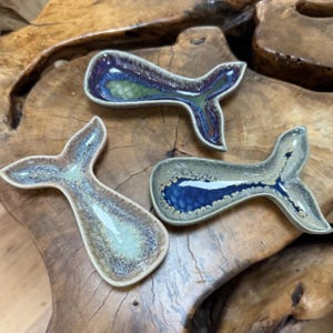 May include: Three ceramic whale tail-shaped dishes with a glossy finish. The dishes feature a variety of colours, including blue, green, and brown, with a speckled or marbled effect. The dishes are displayed on a wooden surface.