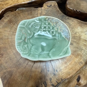 May include: A light green ceramic dish with an elephant design. The dish has a textured surface and a slightly raised rim. The elephant is depicted in a detailed relief, with intricate patterns. The rim has a speckled, brown and gold glaze.