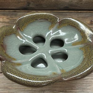 May include: A ceramic, flower-shaped dish with a light blue interior and a speckled, brown-toned rim. The dish has five circular openings in the center. The glaze has a crackle effect.