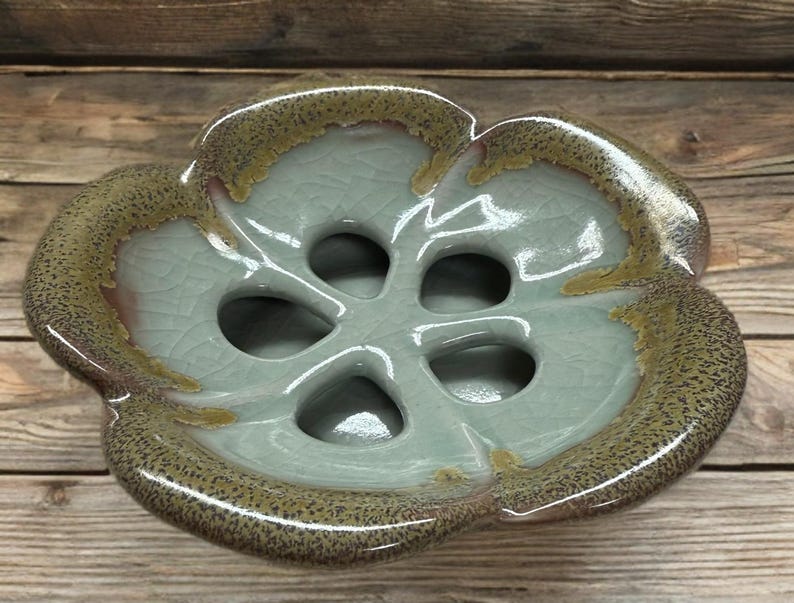 May include: A ceramic, flower-shaped dish with a light blue interior and a speckled, brown-toned rim. The dish has five circular openings in the center. The glaze has a crackle effect.