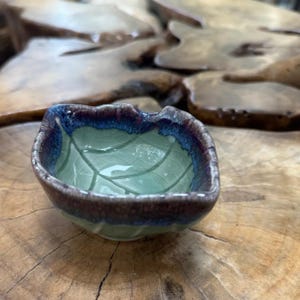 May include: A small, leaf-shaped ceramic bowl with a light green interior and a dark blue and brown rim. The bowl features a detailed leaf vein pattern and is set on a wooden surface.