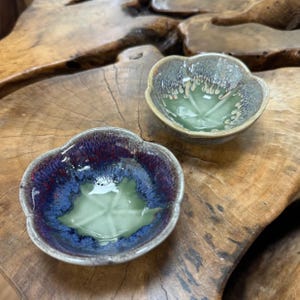 May include: Two small, ceramic bowls with a floral shape. One bowl has a deep blue and purple rim with a green interior. The other bowl has a grey rim and a green interior. Both bowls are on a wooden surface.
