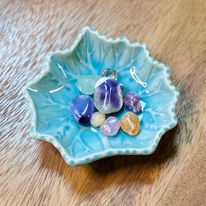 May include: A teal blue ceramic dish shaped like a leaf with a scalloped edge, holding a collection of small, colourful gemstones. The gemstones include amethyst, clear quartz, rose quartz, and citrine.