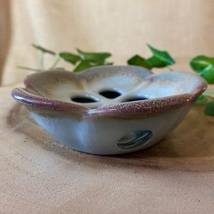 May include: A ceramic, flower-shaped bowl with a light blue base, a reddish-brown rim, and three holes in the center. The bowl has a glossy finish and is likely handmade. The background is a neutral tan color.