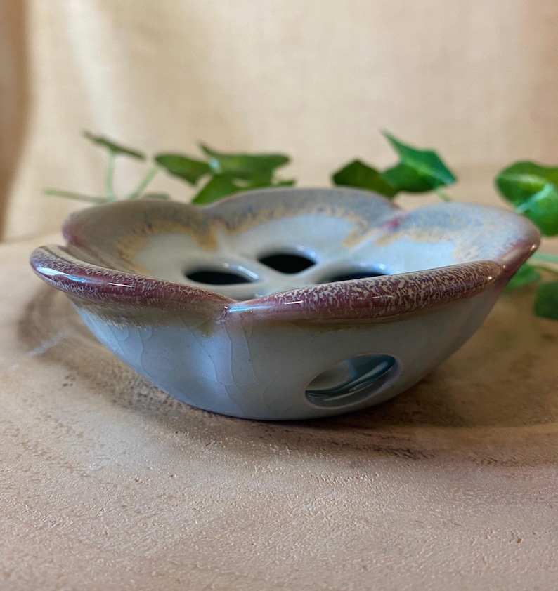 May include: A ceramic, flower-shaped bowl with a light blue base, a reddish-brown rim, and three holes in the center. The bowl has a glossy finish and is likely handmade. The background is a neutral tan color.