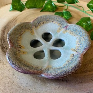 May include: A ceramic soap dish with a flower-like design. The dish has a light blue center with a crackle glaze and a rim that transitions from purple to gold. The dish has a floral shape with five openings.