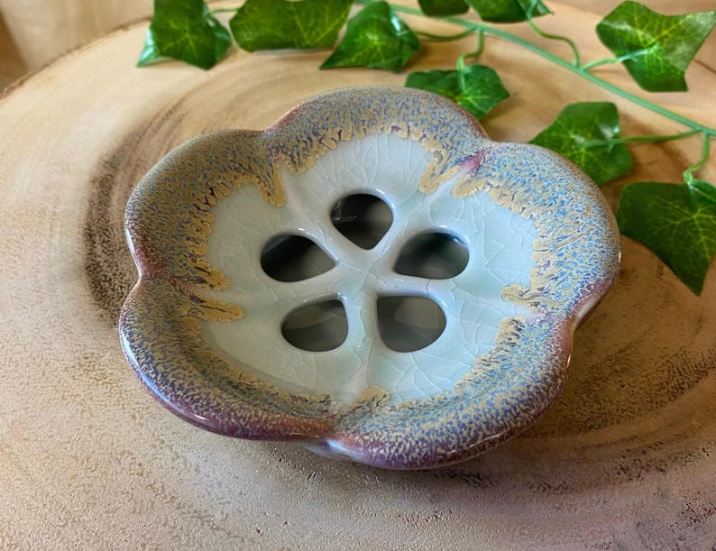 May include: A ceramic soap dish with a flower-like design. The dish has a light blue center with a crackle glaze and a rim that transitions from purple to gold. The dish has a floral shape with five openings.