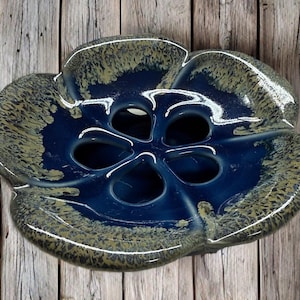 May include: A ceramic, flower-shaped bowl with a deep blue interior and a speckled, olive-green and blue exterior. The bowl has five openings in the center and is likely used for decorative purposes or as a serving dish.