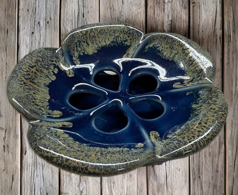 May include: A ceramic, flower-shaped bowl with a deep blue interior and a speckled, olive-green and blue exterior. The bowl has five openings in the center and is likely used for decorative purposes or as a serving dish.
