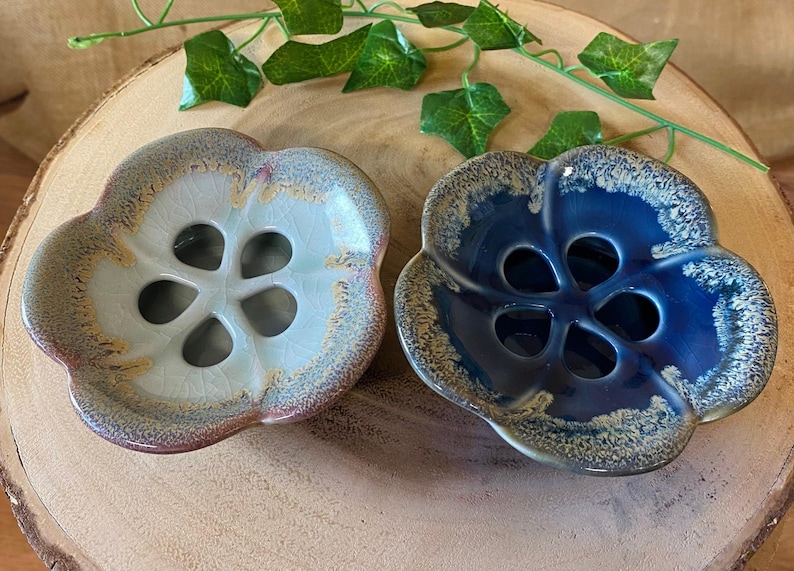 May include: Two ceramic flower-shaped dishes. One is light blue with a brown rim, the other is dark blue with a gold rim. Both have a central floral design with four openings. Green leaves are in the background.