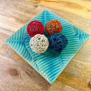 May include: Four decorative balls made of woven material in red, orange, white, and blue colours are arranged on a square, turquoise ceramic tray with a leaf design.