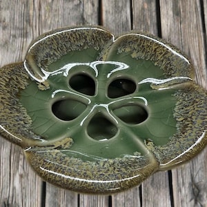 May include: A ceramic flower-shaped vase in shades of green and brown. The vase has a glossy finish and a unique design with multiple openings in the center. The edges have a textured, speckled appearance, adding visual interest to the piece.