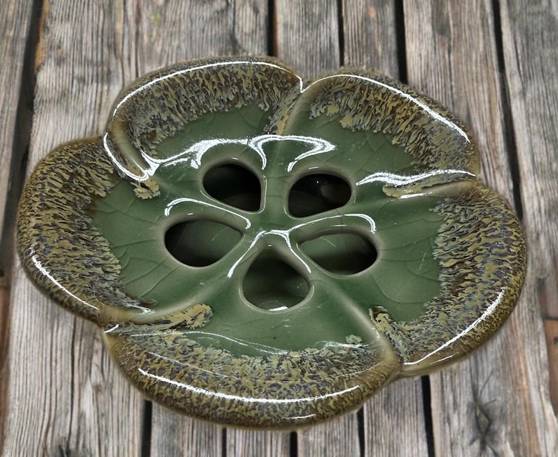 May include: A ceramic flower-shaped vase in shades of green and brown. The vase has a glossy finish and a unique design with multiple openings in the center. The edges have a textured, speckled appearance, adding visual interest to the piece.