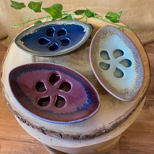 May include: Three ceramic flower frog vases in blue, burgundy, and pale green, each with a unique speckled rim. The oval-shaped vases have a floral design with multiple holes. They are arranged on a wooden surface.