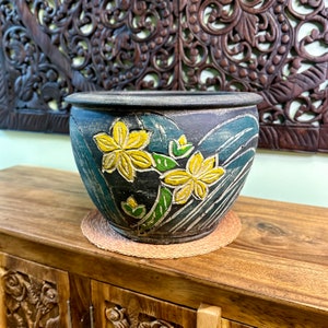 May include: A dark blue ceramic plant pot with yellow and green floral patterns. The pot is round with a wide opening and rests on a woven mat. The background includes a wooden cabinet and carved wall decor.