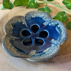 May include: A blue ceramic flower-shaped dish with five holes in the center. The dish has a glossy finish with a white and tan speckled rim. The dish is on a wooden surface with green leaves in the background.