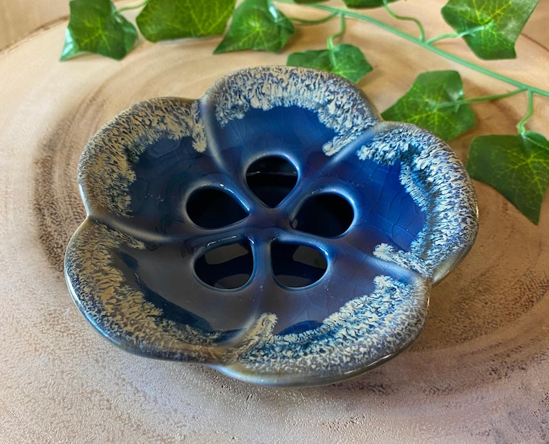 May include: A blue ceramic flower-shaped dish with five holes in the center. The dish has a glossy finish with a white and tan speckled rim. The dish is on a wooden surface with green leaves in the background.