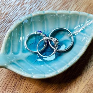 May include: A light blue ceramic leaf-shaped dish with three silver rings with blue gemstones.