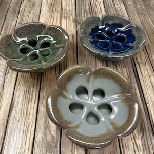 May include: Three ceramic flower-shaped bowls in shades of green, blue, and off-white. Each bowl has a central design with multiple holes. The bowls have a glossy finish and sit on a wooden surface.