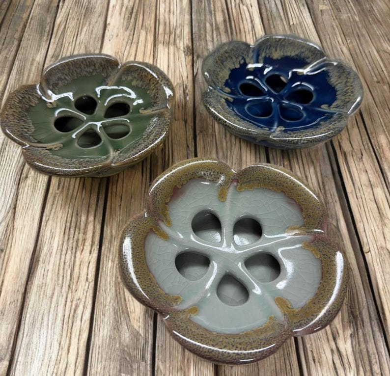 May include: Three ceramic flower-shaped bowls in shades of green, blue, and off-white. Each bowl has a central design with multiple holes. The bowls have a glossy finish and sit on a wooden surface.