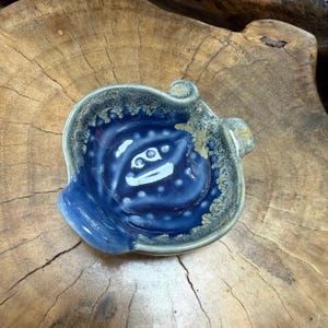 May include: A handcrafted ceramic dish with an unusual, natural form. The dish has a deep blue glaze with lighter blue and beige accents around the edge and inside. The design includes a central spiral pattern with white dots.