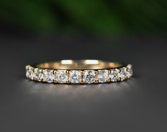 Può includere: Un anello a fascia in oro con numerosi piccoli diamanti a taglio quadrato. L'anello è esposto su una superficie scura, con uno sfondo verde sfocato. I diamanti brillano.
