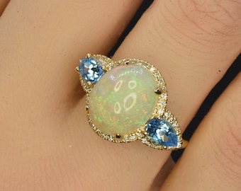 May include: A gold ring with a large white opal gemstone surrounded by small diamonds. Two smaller blue gemstones are set on either side of the opal.