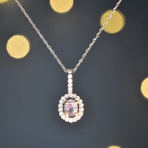 Puede incluir: Un delicado collar de cadena dorada con un colgante que presenta una piedra ovalada de color rosa claro rodeada por un halo de piedras transparentes m&aacute;s peque&ntilde;as.