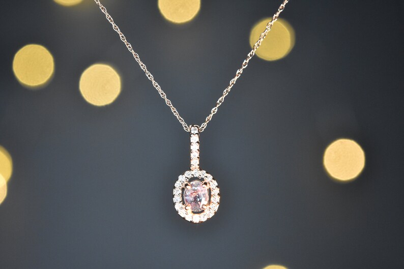 Puede incluir: Un delicado collar de cadena dorada con un colgante que presenta una piedra ovalada de color rosa claro rodeada por un halo de piedras transparentes m&aacute;s peque&ntilde;as.