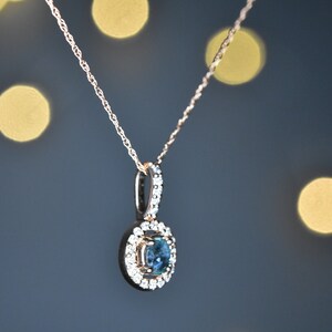 Puede incluir: Un delicado collar de cadena de oro con un colgante que presenta una piedra preciosa azul redonda rodeada por un halo de piedras preciosas transparentes m&aacute;s peque&ntilde;as.