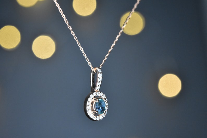 Puede incluir: Un delicado collar de cadena de oro con un colgante que presenta una piedra preciosa azul redonda rodeada por un halo de piedras preciosas transparentes m&aacute;s peque&ntilde;as.