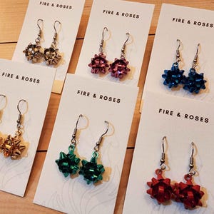 May include: Earrings in various colors, including silver, pink, blue, gold, green, and red, are displayed on white cards. Each pair of earrings features a bow design and is labeled with the text "FIRE & ROSES".