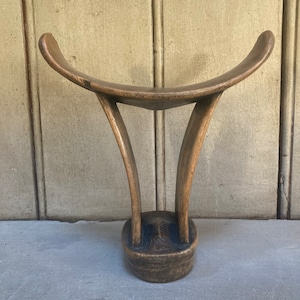 May include: A wooden headrest carved in the shape of a crescent moon with two curved supports. The headrest is dark brown and has a smooth, polished finish.