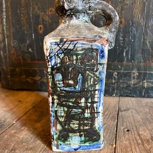Może przedstawiać: Ceramiczny dzbanek o kwadratowym korpusie i uchwycie. Dzbanek jest ozdobiony abstrakcyjnymi czarnymi, zielonymi, niebieskimi i brązowymi liniami i kształtami.