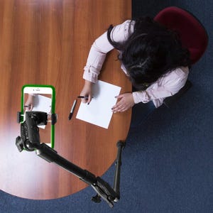Może przedstawiać: Czarny, regulowany wysięgnik z podstawą zaciskową i uchwytem na smartfon w zielonej obudowie. Uchwyt jest ustawiony tak, aby trzymać smartfon bezpośrednio nad powierzchnią drewnianego biurka, umożliwiając nagrywanie dokumentów i pisania.