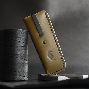 May include: Olive green leather case with a black metal clip and a debossed bird logo. A spool of black thread and small silver objects are next to the case. The case is angled against a dark background.