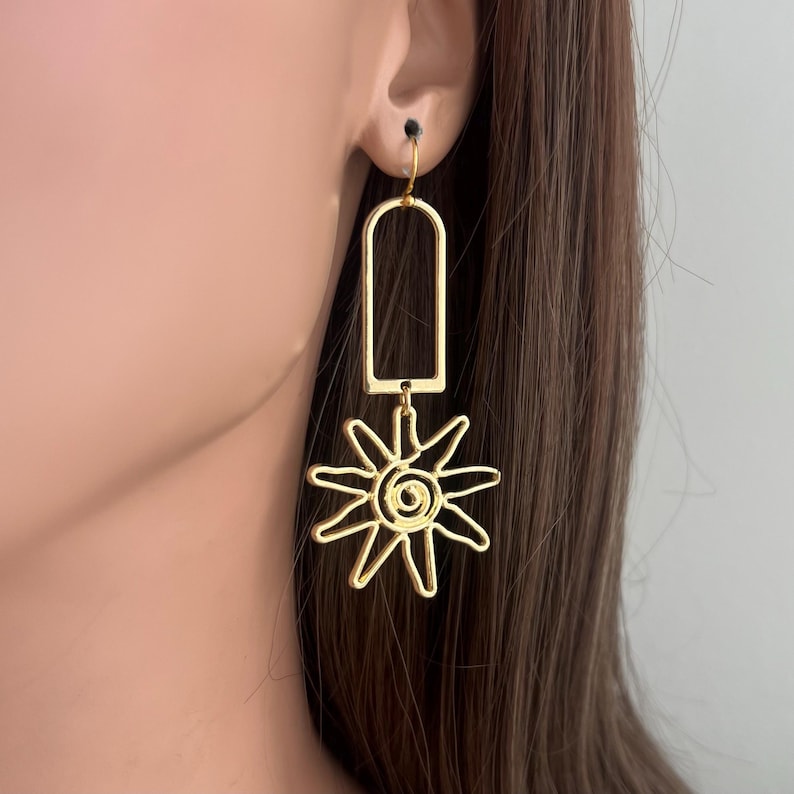 May include: Gold-toned dangle earrings featuring a geometric rectangle shape with a sun design hanging from it.