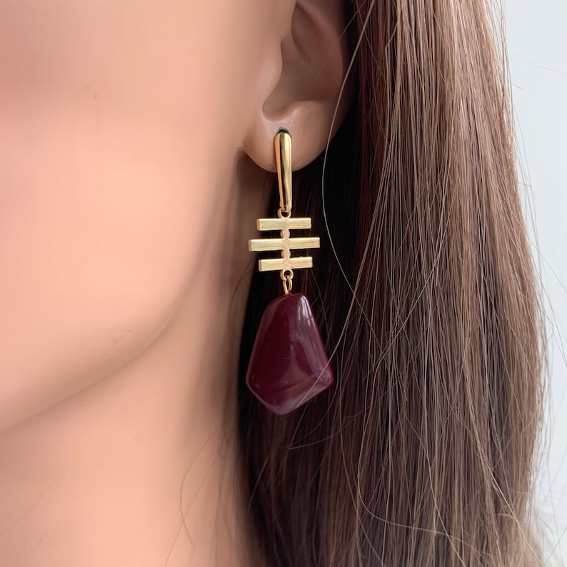 May include: Gold geometric dangle earrings with a burgundy teardrop-shaped stone.