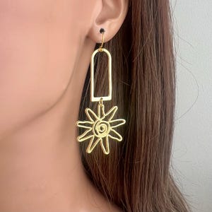 May include: Gold dangle earrings with a sun-shaped charm. The sun charm has a spiral design in the center.