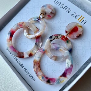 May include: A pair of multi-colored, round, acrylic earrings with a gold-tone hook. The earrings feature three interlocking circles, each with a different pattern. The earrings are in a white box with the text "URBAN ZELIA" and a website address.