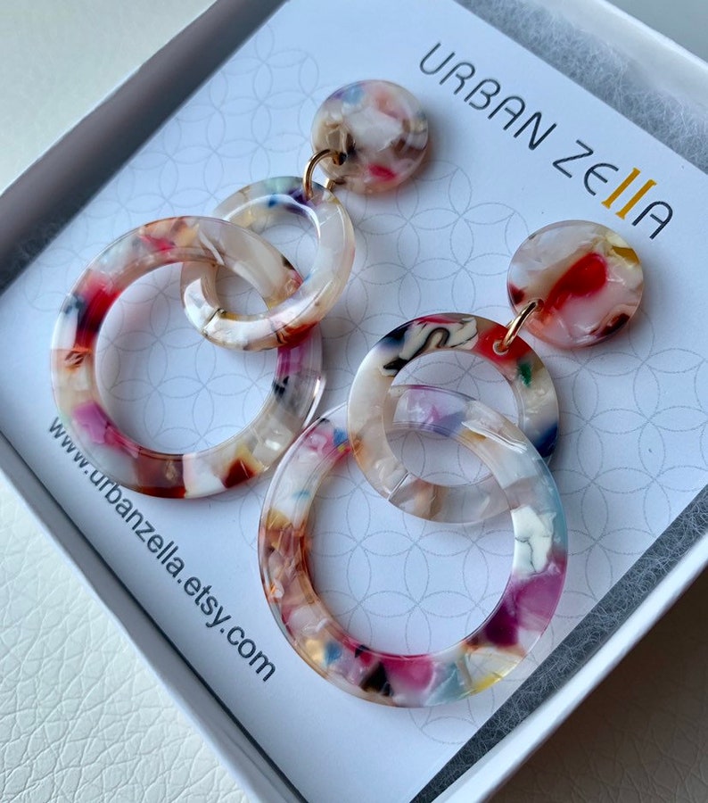 May include: A pair of multi-colored, round, acrylic earrings with a gold-tone hook. The earrings feature three interlocking circles, each with a different pattern. The earrings are in a white box with the text "URBAN ZELIA" and a website address.
